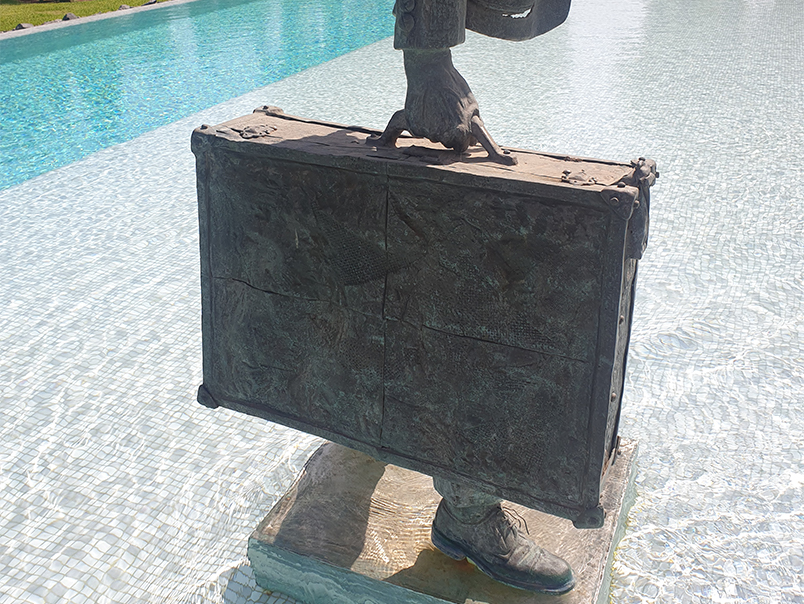 Gros plan sur la valise en bronze patiné portée par le personnage de la Sculpture Peigne